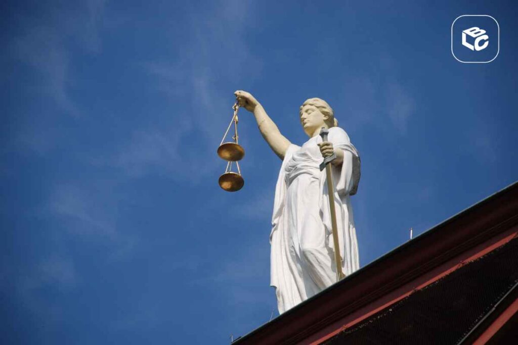 Estátua branca da Justiça vista de baixo, segurando uma balança dourada erguida em uma das mãos e uma espada na outra. A escultura está posicionada sobre o topo de um edifício, com céu azul limpo ao fundo. No canto superior direito, há um logotipo branco dentro de um quadrado arredondado.