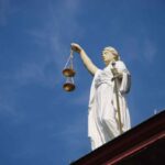 Estátua branca da Justiça vista de baixo, segurando uma balança dourada erguida em uma das mãos e uma espada na outra. A escultura está posicionada sobre o topo de um edifício, com céu azul limpo ao fundo. No canto superior direito, há um logotipo branco dentro de um quadrado arredondado.