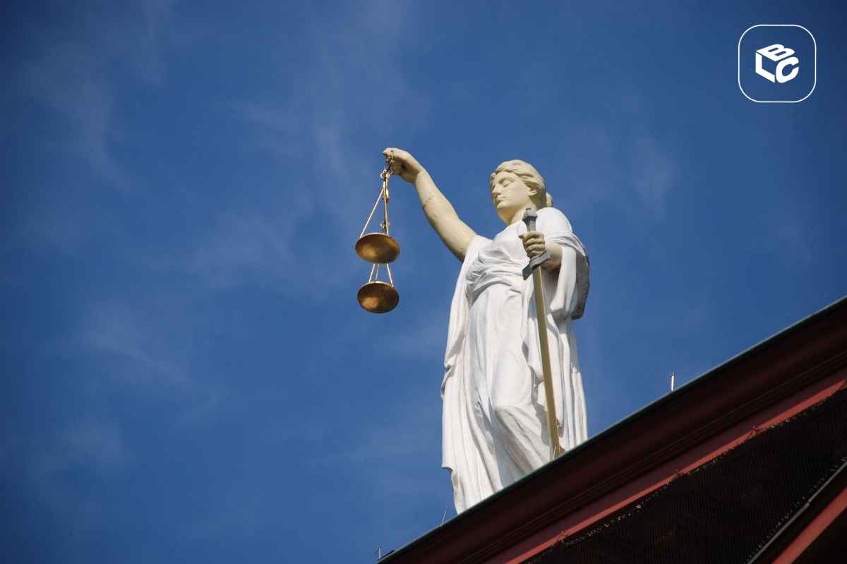 Estátua branca da Justiça vista de baixo, segurando uma balança dourada erguida em uma das mãos e uma espada na outra. A escultura está posicionada sobre o topo de um edifício, com céu azul limpo ao fundo. No canto superior direito, há um logotipo branco dentro de um quadrado arredondado.